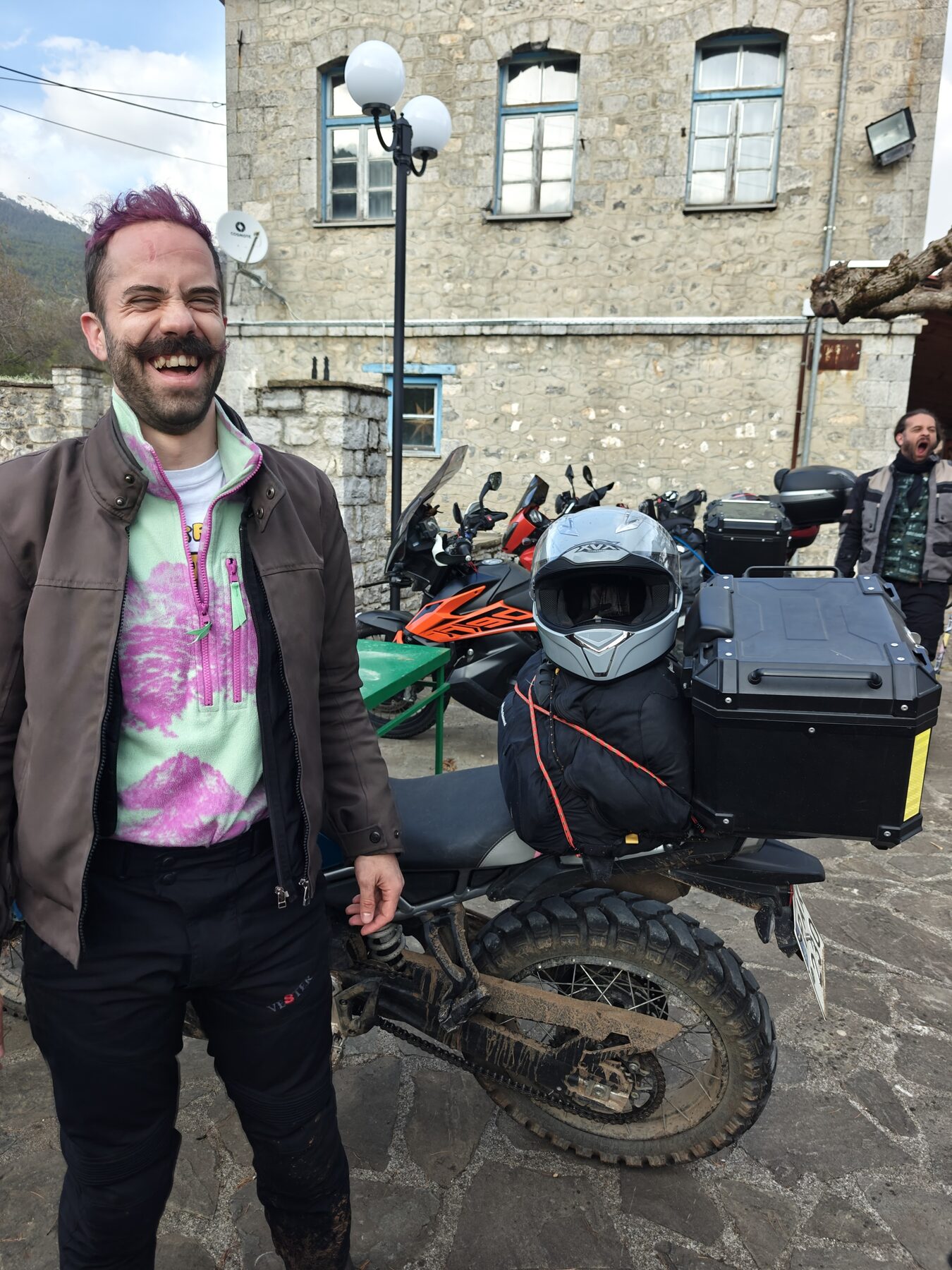 Orfeas laughing beside a muddy motorbike in a mountain village