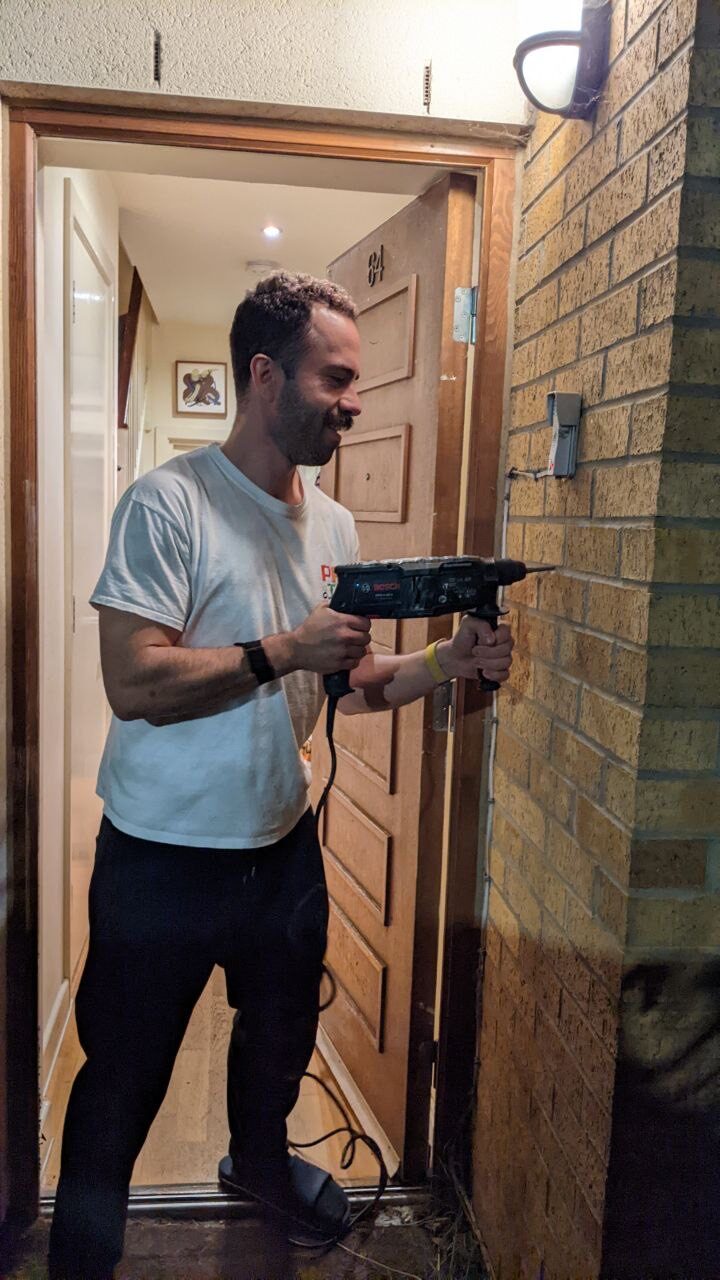Orfeas using an electric drill beside a front door