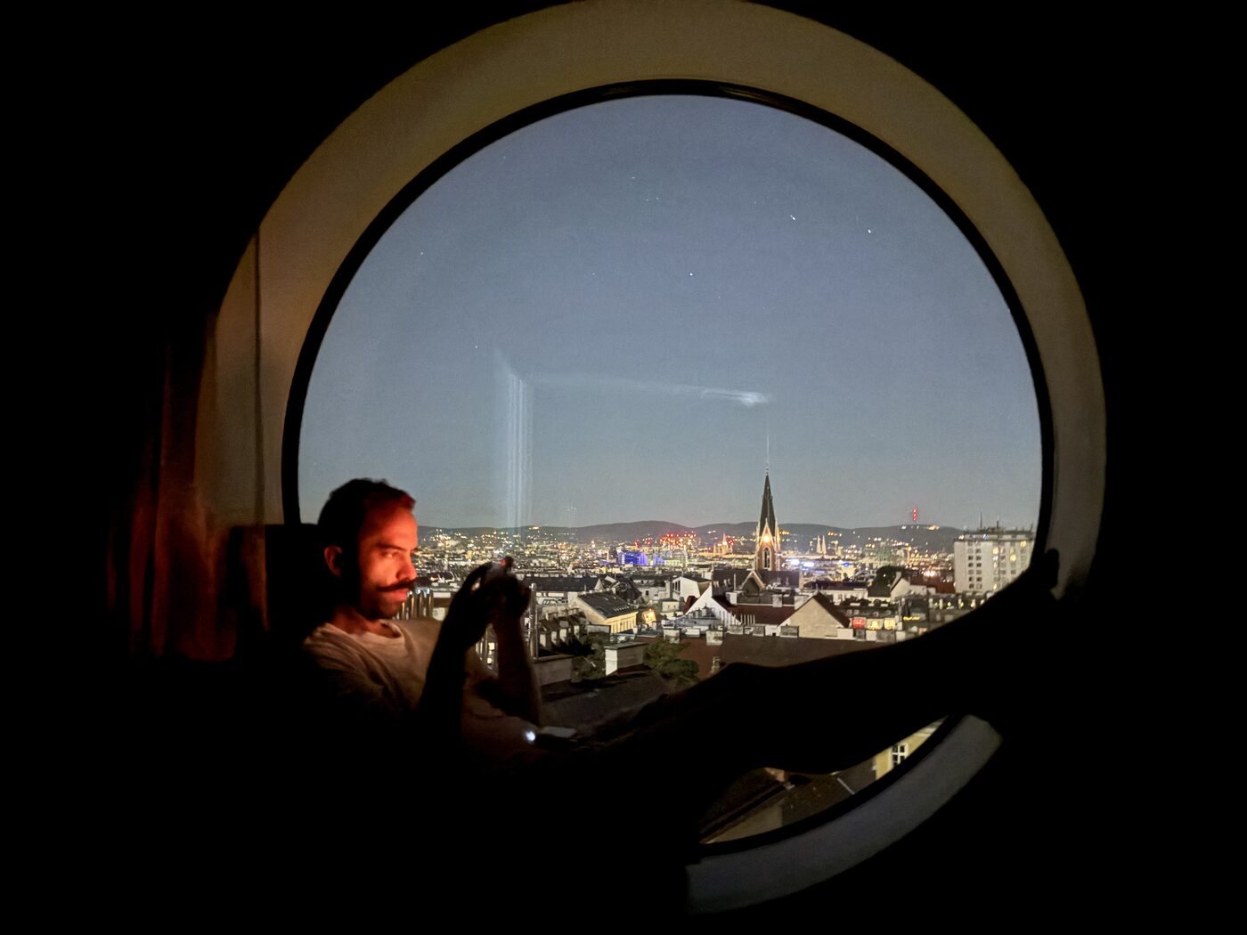 Orfeas relaxing beside a round window overlooking city lights at night